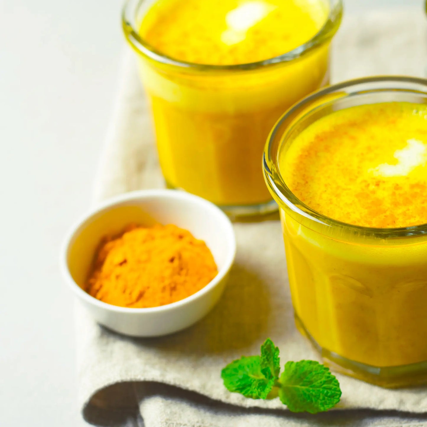 Two glasses of yellow turmeric latte with a small bowl of turmeric powder and mint leaves on a light background.